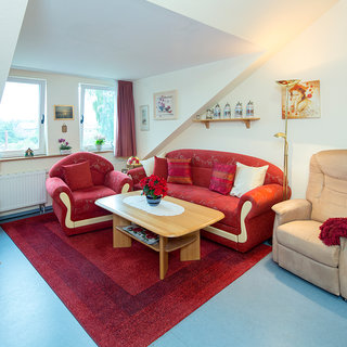 Das Bild zeigt ein gemütliches Wohnzimmer mit einem roten Sofa und zwei Sesseln. In der Mitte steht ein Holztisch mit einer Blumenvase. Der Raum hat einen Teppich, große Fenster und ist hell erleuchtet. Es gibt auch einen Fernseher und einige dekorative Elemente an den Wänden.