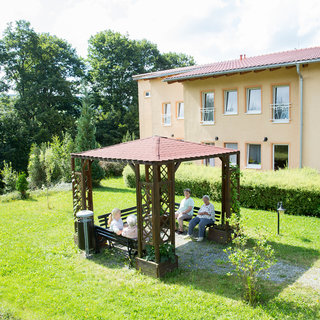 Eine Gruppe von Menschen sitzt unter einem Pavillon in einem Garten. Im Hintergrund ist ein Gebäude sichtbar, umgeben von Bäumen und grüner Wiese. Die Atmosphäre wirkt ruhig und einladend.