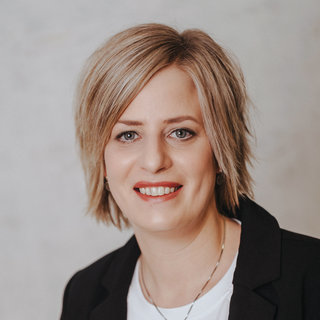 Das Bild zeigt eine lächelnde Frau mit kurzen, blonden Haaren, die einen schwarzen Blazer und ein weißes T-Shirt trägt. Der Hintergrund ist neutral und hell. Ihre Ausstrahlung ist freundlich und professionell.