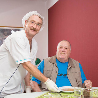 Ein Pfleger in uniform mit Haube lächelt, während er einem älteren Mann, der am Tisch sitzt, eine Schüssel Suppe serviert. Der Mann wirkt zufrieden und trägt ein blaues T-Shirt und eine graue Weste. Im Hintergrund sind unauffällige Wohnräume sichtbar.