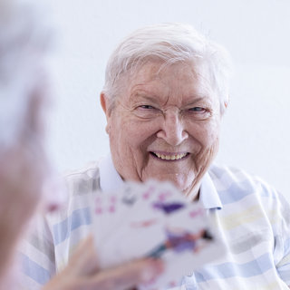 Eine ältere Person mit grauen Haaren lächelt fröhlich, während sie Karten hält. Im Hintergrund ist eine weitere Person anscheinend ebenfalls mit Karten beschäftigt. Der Ausdruck der ersten Person vermittelt Freude und Geselligkeit beim Spielen.