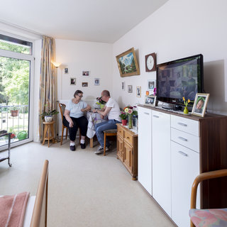 In einem hellen Wohnzimmer sitzen drei Personen zusammen. Eine Frau hilft einem älteren Mann, während eine andere Frau lächelt. Im Hintergrund sind Pflanzen und Fotos an der Wand sichtbar, und ein Fernseher steht auf einem Möbelstück. Licht strömt durch große Fenster.