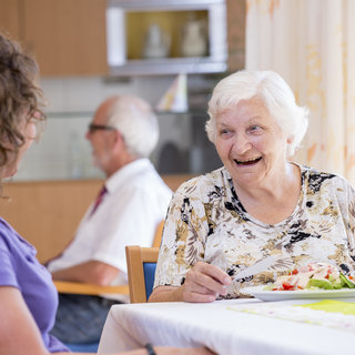 Eine ältere Frauen sitzt lächelnd am Tisch und genießt ihre Mahlzeit. Sie unterhält sich mit einer jüngeren Frau, die ihr gegenüber sitzt. Im Hintergrund ist ein weiterer älterer Mann zu sehen. Das Ambiente wirkt freundlich und einladend.