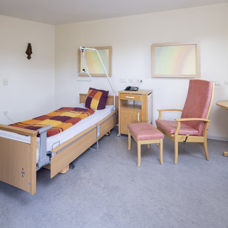 Ein helles Zimmer mit einem Pflegebett, das mit einer gestreiften Decke und Kissen ausgestattet ist. Neben dem Bett steht ein Tisch mit einer Lampe und einer weiteren Sitzgelegenheit. An der Wand hängen zwei gemalte Bilder. Der Raum wirkt freundlich und einladend.