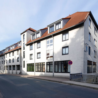 Das Bild zeigt ein modernes, mehrstöckiges Wohngebäude an einer ruhigen Straße. Es hat mehrere Fenster und ein Satteldach. Die Fassade ist hell gestrichen, und ein grüner Eingang ist sichtbar. Im Vordergrund verläuft ein gepflasterter Weg.
