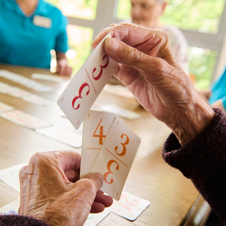 Eine ältere Person hält Karten mit den Zahlen 3, 4 und 3 in der Hand. Im Hintergrund sind weitere Personen zu sehen, die sich mit dem Kartenspiel beschäftigen. Der Tisch ist mit weiteren Karten bedeckt, und es gibt eine helle, freundliche Umgebung.