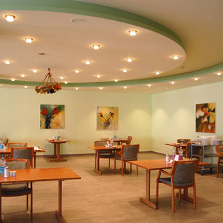 Ein heller Restaurantraum mit Holztischen und Stühlen. An den Wänden hängen mehrere Gemälde mit bunten Motiven. Die Beleuchtung ist freundlich und sorgt für eine einladende Atmosphäre. Die Gaststättenmöbel sind ordentlich angeordnet und laden zum Verweilen ein.