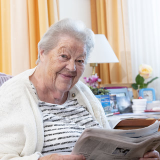 Eine ältere Frau sitzt auf einem Sessel und lächelt, während sie eine Zeitung in der Hand hält. Sie trägt einen hellen Pulli und eine Strickjacke. Im Hintergrund sind Tageslichter und eine Zimmerpflanze zu sehen.