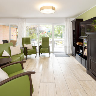 Ein heller Wohnraum mit großen Fenstern, die auf einen Gartenblick zeigen. Drei grüne Sessel mit weißen Kissen stehen um kleine Tische. An einer Wand steht ein schwarzer Schrank mit Pflanzen und Regal. Helle Fliesen schaffen eine freundliche Atmosphäre.