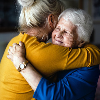 Zwei Frauen umarmen sich herzlich. Eine hat lange blonde Haare und trägt ein gelbes Oberteil, die andere hat graue Haare und trägt ein blaues Oberteil. Beide zeigen ein Lächeln, das Zuneigung und Freude ausdrückt.