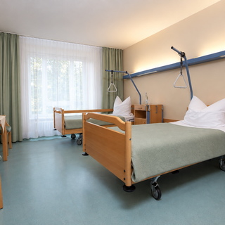 Ein helles Krankenzimmer mit zwei verstellbaren Krankenhausbetten, einem Tisch und Stühlen. Vor einem Fenster mit Vorhängen steht eine Vase mit Blumen. Der Boden ist aus strapazierfähigem Material, und die Wände sind in sanften Farben gehalten.