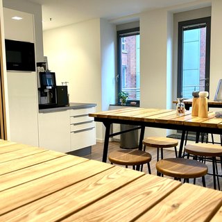 Eine helle Küche mit Holztischen und Stühlen. Im Hintergrund steht ein Kaffeeautomat und große Fenster lassen Tageslicht herein. Der Raum wirkt modern und einladend.