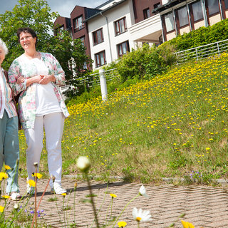 Zwei Frauen stehen auf einem Weg, umgeben von einer bunten Wiese mit gelben Blumen. Die ältere Frau trägt eine Braune Jacke, die jüngere Frau ein weißes Oberteil mit einer bunten Jacke. Im Hintergrund sind mehrstöckige Gebäude zu sehen. Es ist ein sonniger Tag.