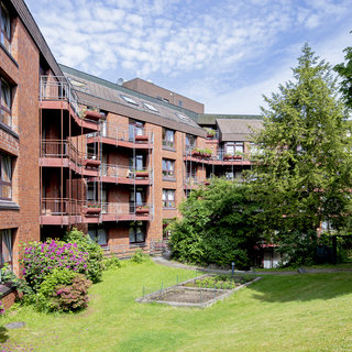 Eine Ansicht eines Mehrfamilienhauses mit roten Backsteinen. Einige Balkone sind sichtbar, auf denen Pflanzen stehen. Im Vordergrund gibt es einen kleinen Garten mit Grünflächen und Büschen, umgeben von Bäumen. Der Himmel ist blau mit etwas Bewölkung.