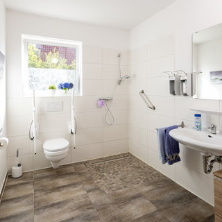 Ein modernes Badezimmer mit weißen Fliesen und einem großen Fenster. Es gibt eine Dusche mit Haltegriffen, eine Toilette mit Haltegriffen, ein Waschbecken mit einem Handtuch und Pflegeprodukten. Holz- und graue Fliesen sorgen für eine schlichte, einladende Atmosphäre. Blumen auf dem Fensterbrett runden die Einrichtung ab.