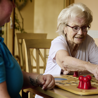 Eine ältere Frau mit Brille sitzt an einem Tisch und spielt mit bunten Spielfiguren, während eine jüngere Person in blauer Kleidung sie beobachtet. Die Atmosphäre ist freundlich und entspannt.