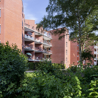 Ein Mehrfamilienhaus aus roten Ziegeln mit mehreren Balkonen. Grüne Sträucher und Bäume umgeben das Gebäude. Der Himmel ist blau und es ist eine sonnige Stimmung wahrzunehmen.
