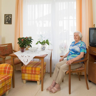 Eine ältere Frau sitzt in einem gemütlichen Wohnzimmer an einem Tisch mit Blumen. Der Raum hat helle Vorhänge und eine warme Atmosphäre. Im Hintergrund sind ein Fernseher und einige Bilder an der Wand zu sehen.