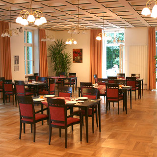 Ein einladender Speisesaal mit mehreren Tischen und Stühlen. Die Tische sind gedeckt und in der Mitte des Raumes stehen Pflanzen. Helle Fenster sorgen für eine freundliche Atmosphäre, während die Wände mit orangefarbenen Vorhängen und einem Bild dekoriert sind.