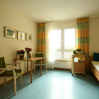 Das Bild zeigt ein helles Zimmer mit einem Fenster, durch das Tageslicht fällt. Es gibt einen Bett, einen Tisch mit Stühlen und eine kleine Kommode. An der Wand hängen Bilder, und auf dem Tisch stehen Blumen. Die Farben der Vorhänge sind gelb und grün.