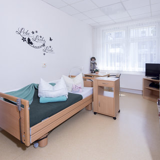 Ein Krankenhauszimmer mit einem verstellbaren Bett, das mit Kissen und einer Decke ausgestattet ist. Am Fenster steht ein kleiner Tisch mit einem Stuhl. An der Wand sind dekorative Elemente angebracht, und ein Fernseher sowie ein kleiner Schrank sind vorhanden.