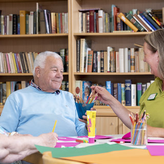 Eine ältere Frau und ein älterer Mann sitzen an einem Tisch, während sie kreativ arbeiten. Die Frau reicht dem Mann eine Schüssel mit Stiften. Im Hintergrund sind Bücherregale mit einer Vielzahl von Büchern sichtbar. Der Raum wirkt freundlich und einladend.