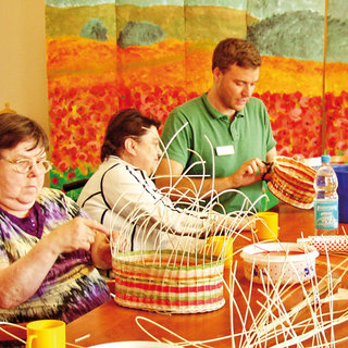Drei Personen sitzen an einem Tisch und werken an der Korbflechterei. Die Männer und Frauen sind konzentriert bei der Arbeit, während sie Materialien in bunten Tönen verwenden. Im Hintergrund ist eine leuchtende Wandmalerei zu sehen, die eine blühende Landschaft darstellt.