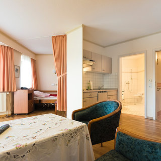 Das Bild zeigt einen hellen Wohnraum mit einer Sitzecke, einem Tisch und einer offenen Küchenzeile. Im Hintergrund ist ein Schlafzimmer mit einem Bett und ein Badezimmer mit Tür zu sehen. Große Fenster lassen Tageslicht herein. Die Einrichtung ist funktional und gemütlich.