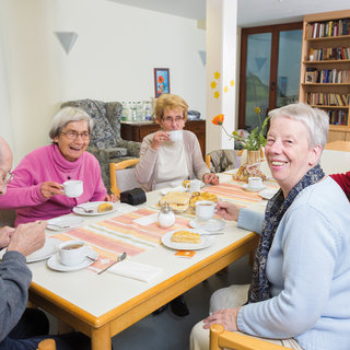 Eine Gruppe älterer Menschen sitzt um einen Tisch und genießt Kaffee und Gebäck. Sie wirken freundlich und lächeln, während sie miteinander sprechen. Im Hintergrund sind Bücherregale zu sehen, die eine einladende Atmosphäre schaffen.