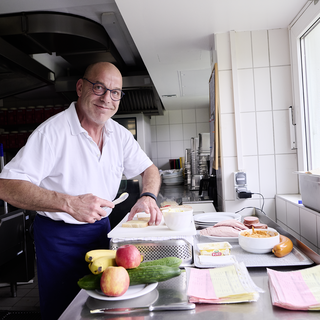Ein Koch steht in einer gut ausgestatteten Küche an einem Tisch. Er lächelt und schneidet Gemüse. Auf dem Tisch liegen verschiedene Lebensmittel wie Äpfel, Bananen und Gurken sowie einige Teller und Unterlagen.
