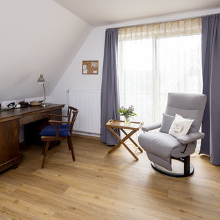 Ein einladender Raum mit einem Holztisch, einem blauen Stuhl und einer Leselampe. Neben dem Tisch steht ein ruhiger Sessel, während ein Holztisch mit einer Vase und Blumen darauf daneben steht. Das Licht strömt durch große Fenster mit transparenten Vorhängen.