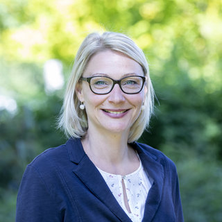 Das Bild zeigt eine lächelnde Frau mit blonden, schulterlangen Haaren und Brille. Sie trägt eine dunkle Jacke und ein helles Oberteil. Der Hintergrund ist unscharf und zeigt grüne Pflanzen.