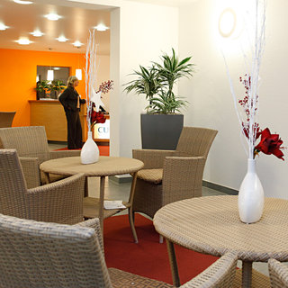 Der Raum zeigt eine gemütliche Sitzecke mit vier geflochtenen Stühlen und einem runden Tisch. In der Mitte steht eine Vase mit dekorativen Blumen. Im Hintergrund sind Pflanzen und ein Empfangstresen sichtbar, während eine Person im Vordergrund steht.