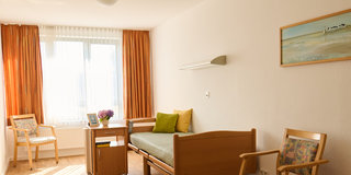 Ein helles Zimmer mit Holzboden, das eine wütende Sitzecke mit zwei Stühlen und einem Bett zeigt. Das Bett hat eine grüne Decke und ein Kissen. Neben dem Bett steht ein kleiner Tisch mit einem Blumengesteck. Große Fenster lassen viel Tageslicht herein.