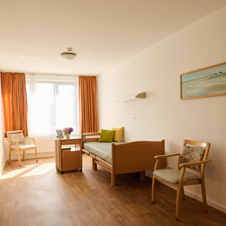 Ein helles Zimmer mit Holzboden, das eine wütende Sitzecke mit zwei Stühlen und einem Bett zeigt. Das Bett hat eine grüne Decke und ein Kissen. Neben dem Bett steht ein kleiner Tisch mit einem Blumengesteck. Große Fenster lassen viel Tageslicht herein.