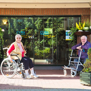 Zwei ältere Menschen sitzen vor einem Gebäude. Eine Person sitzt im Rollstuhl, die andere benutzt einen Gehwagen. Beide sehen freundlich aus und lächeln in die Kamera. Im Hintergrund sind Pflanzen und das Schild "CURA" zu sehen.