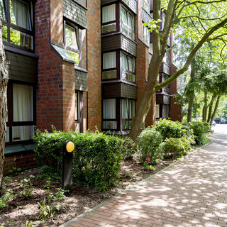 Ein gepflasterter Weg verläuft an einem modernen, roten Ziegelgebäude. Im Vordergrund stehen Bäume und Sträucher mit bunten Blumen. Zwei Personen spazieren auf dem Weg, während Autos am Ende des Weges geparkt sind. Die Szene ist hell erleuchtet von Tageslicht.