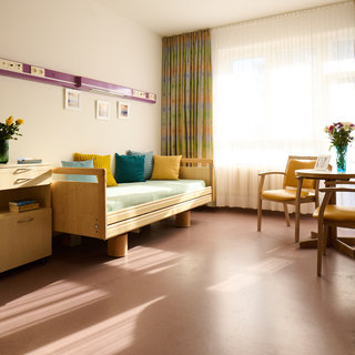 Ein helles Krankenzimmer mit einem einzelnen Bett, zwei Stühlen und einem kleinen Tisch. Auf dem Bett liegen bunte Kissen, und ein Blumenstrauß steht auf dem Tisch. Fenster mit Vorhängen lassen viel Licht hinein, wodurch der Raum freundlich wirkt.