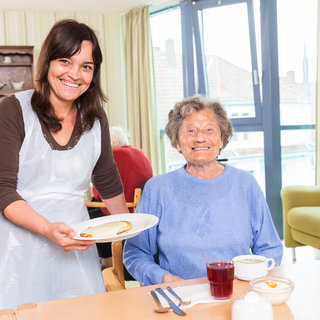 Eine lächelnde Servicekraft reicht einem älteren Menschen in einem Restaurant einen Teller mit Essen. Der ältere Mensch trägt einen blauen Pullover und lächelt zurück. Auf dem Tisch stehen ein Glas Saft und eine Tasse. Helle, freundliche Einrichtung im Hintergrund.