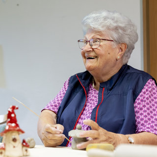 Eine lächelnde ältere Frau sitzt an einem Tisch und gestaltet Keramiken. Sie trägt eine lila Bluse und eine dunkle Weste. In ihrer Hand hält sie ein Werkzeug, während sie konzentriert an einem Kunstwerk arbeitet. Auf dem Tisch sind mehrere Kunstobjekte zu sehen.