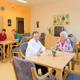 In einem hellen Aufenthaltsraum spielen zwei Senioren, ein Mann und eine Frau, am Tisch Karten. Im Hintergrund sind zwei jüngere Männer, die ebenfalls an einem Spiel beteiligt sind. Die Atmosphäre wirkt freundlich und gesellig, mit Pflanzen und Wanddekorationen im Raum.