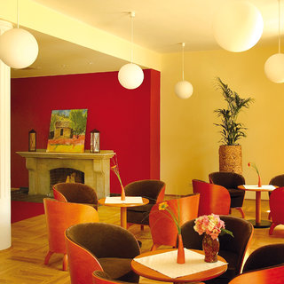 Ein eleganter Raum mit orangefarbenen und braunen Sesseln um runde Tische. Im Hintergrund ist ein Kamin und ein Gemälde zu sehen. Die Wände sind in sanften Farben gehalten, und große, runde Lampen hängen von der Decke. Eine Pflanze in einem Topf ergänzt die Atmosphäre.