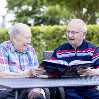 Zwei ältere Männer sitzen an einem Tisch im Freien und schauen gemeinsam in ein Buch. Beide lachen und scheinen eine angenehme Zeit miteinander zu verbringen. Im Hintergrund sind grüne Bäume zu sehen.