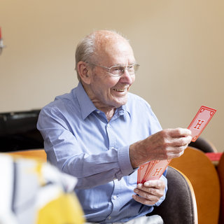 Ein älterer Mann in einem blauen Hemd lächelt, während er rote Spielkarten in der Hand hält. Er sitzt in einem gemütlichen Raum, umgeben von anderen Personen, die nicht im Fokus stehen. Der Mann scheint Freude am Spiel zu haben.
