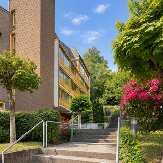 Ein moderner Gebäudekomplex mit gelben Fenstern, umgeben von üppigem Grün und blühenden Pflanzen. Ein Weg mit Treppen führt zur Eingangstür des Gebäudes, das von niedrigen Bäumen und Sträuchern flankiert wird. Der Himmel ist klar und sonnig.