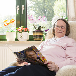 Eine ältere Frau sitzt in einem Sessel und lächelt. Sie trägt einen rosa Pullover und hält eine Zeitschrift in der Hand. Im Hintergrund sind Fenster mit Blumen und Pflanzen sichtbar, die eine freundliche Atmosphäre schaffen.