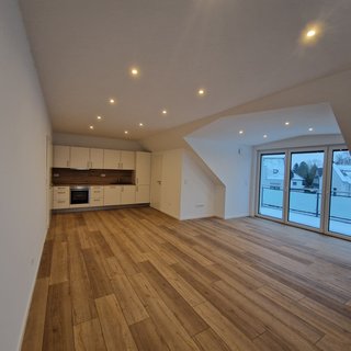 Das Bild zeigt eine moderne, leere Wohnung mit einem offenen Wohnbereich, einer Küchenzeile in hellem Design und einem großen Fenster mit Balkon. Der Boden ist aus Holz und die Decke ist mit mehreren Lichtquellen ausgestattet.