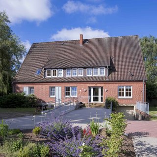 Ein zweigeschossiges, rotes Backsteinhaus mit einem Satteldach. Vor dem Eingang befindet sich eine Rampe. Umgeben von einem gepflegten Garten mit bunten Blumen und Sträuchern. Die Wolken sind leicht und der Himmel ist blau.