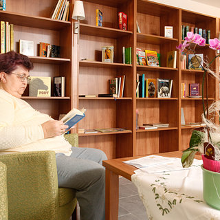Eine ältere Frau sitzt in einem Sessel und liest ein Buch. Hinter ihr befinden sich Regale mit verschiedenen Büchern. Auf dem Tisch vor ihr steht eine Pflanze in einem Topf. Die Atmosphäre wirkt gemütlich und einladend.