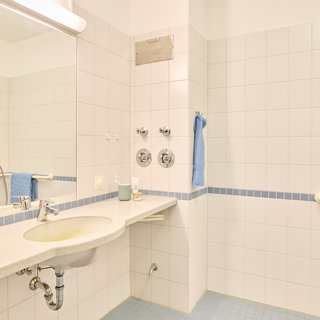 Das Bild zeigt ein modernes Badezimmer mit weißen Fliesen und einem langen Waschbecken. An der Wand befindet sich ein großer Spiegel. Es gibt einen blauen Handtuchhalter und eine Haltegriff für zusätzliche Sicherheit. Das Badezimmer wirkt hell und sauber.